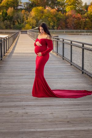 Pregnant woman in elegant red off-shoulder gown stands on wooden boardwalk surrounded by autumn foliage.