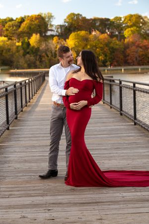 Expecting couple poses on a wooden bridge in autumn, woman wearing an elegant red maternity gown.