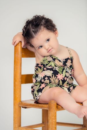 Adorable curly-haired baby girl in a floral swimsuit sits on a wooden chair with a thoughtful expression.