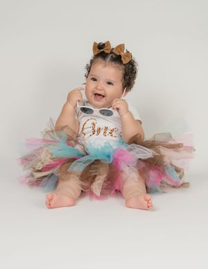 Smiling baby girl in colorful tutu skirt and One shirt with gold bow headband celebrates first birthday.