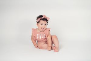 Adorable baby girl in pink One romper with matching bow headband sits on white background.