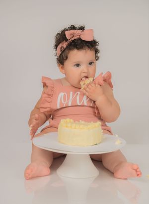 Baby girl in pink ruffle dress eating first birthday smash cake on white cake stand.