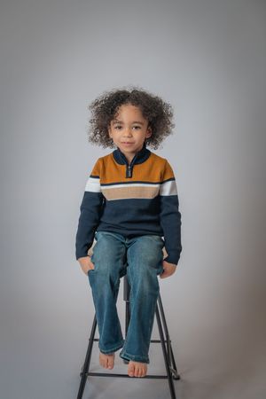 Young child with curly hair sits on stool wearing striped sweater and jeans against gray studio background.