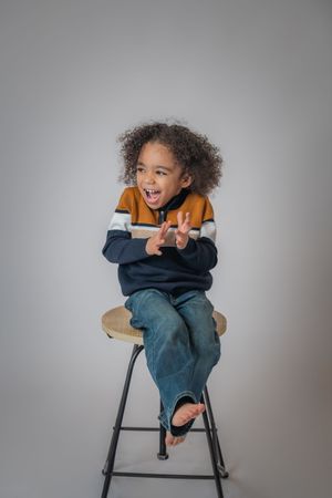 Joyful curly-haired child sitting on a stool making peace signs, wearing a striped sweater and jeans.