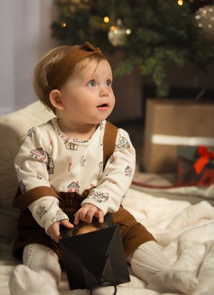 Adorable baby in holiday pajamas sitting near Christmas tree holding a small black gift box.