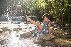 Joyful young woman in floral dress splashing water by a scenic forest waterfall.