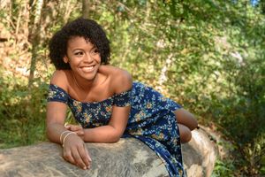 Young Black woman in blue floral dress smiling while relaxing on rocks in nature.