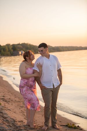 Couple walks barefoot on sandy beach at golden sunset by the water.
