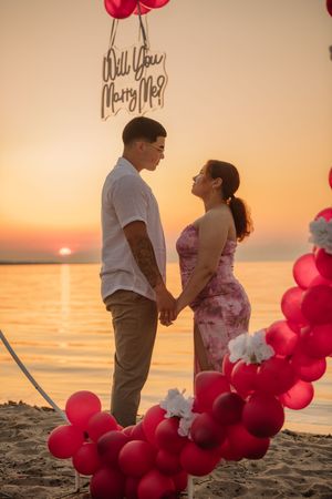 Couple holds hands under Will You Marry Me sign surrounded by red balloons at sunset beach.