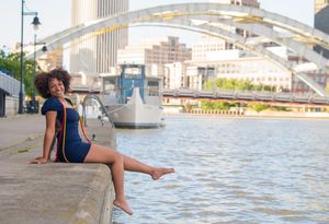 Young Black woman in blue dress sits on waterfront wall with city bridge and river behind her.
