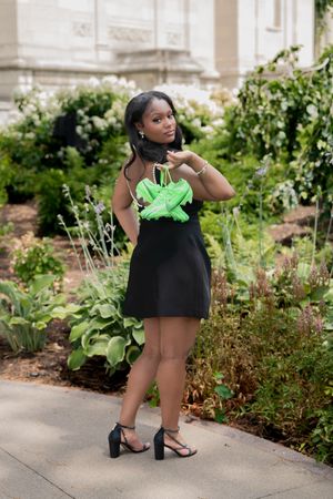 Young woman in black mini dress holding neon green sneakers outdoors near garden background.