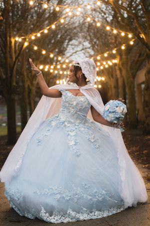 Woman in light blue floral quinceañera ball gown with cape twirling under string lights at dusk.