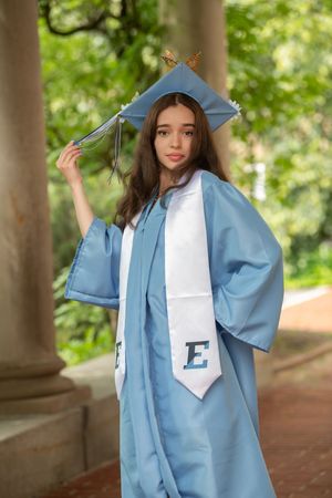 A young woman in a light blue graduation cap and gown with an E stole poses outdoors among columns.