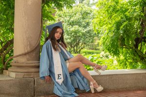 A graduate in blue cap and gown sits relaxed against a stone column in a green outdoor setting.