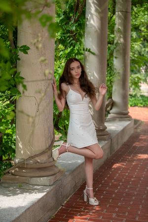 A young woman in a white mini dress poses elegantly between stone columns in a garden walkway.