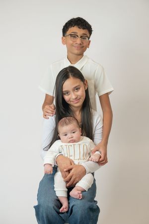 Teenage boy and girl proudly pose with their newborn sibling in a white studio portrait.