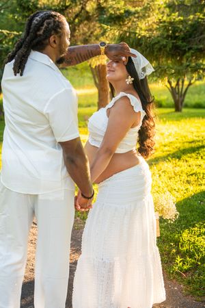 Couple in white outfits holding hands in sunlit park, man gently placing flower in womans hair.