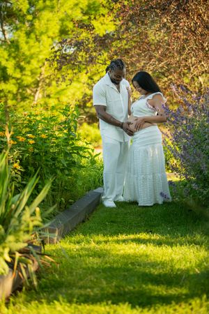 Expecting couple in matching white outfits holding pregnant belly in a lush garden maternity photoshoot.