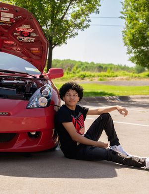 Teen poses beside open hood of red sports car in sunny parking lot.