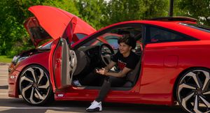 Young man sits in driver seat of red sports car with door open outdoors.