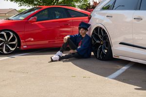 Teen sits between red and white lowered cars in sunny parking lot.