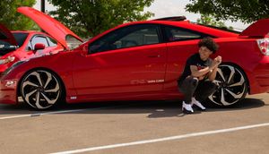 Young man crouches beside lowered red Honda with custom black wheels at car show.