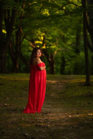 Pregnant woman in flowing red off-shoulder gown stands on a forest path bathed in golden evening light.
