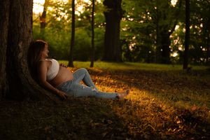 Pregnant woman in white bra and jeans rests against a tree during a golden sunset in the park.
