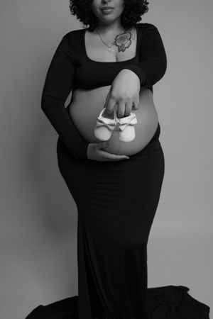 Black and white maternity photo of pregnant woman in black dress holding baby shoes on belly.