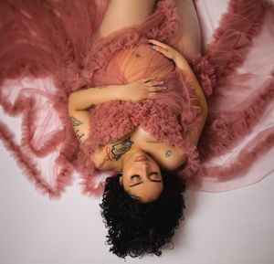 Pregnant woman in pink tulle gown lying down, showing tattoos during artistic maternity photoshoot.