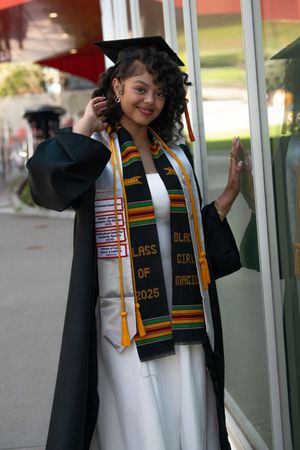 Smiling graduate in cap and gown with kente cloth stole and Class of 2025 sash outdoors.