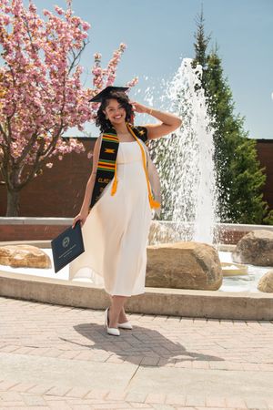 Smiling graduate in white gown and cap with kente stole posing by fountain with cherry blossoms.