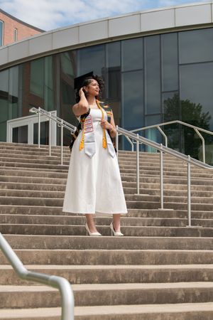 Female graduate in white cap and gown with honor stoles standing on university building steps outdoors.