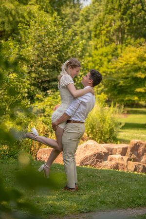 Romantic couple shares a kiss as man lifts woman in a white dress surrounded by lush green park scenery.