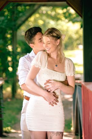 Happy couple embraces under a wooden pergola, woman in white pearl mini dress smiles as partner kisses her cheek.