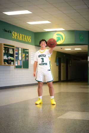 Young basketball player in Greece Olympia Spartans uniform balances a ball on one finger in school hallway.