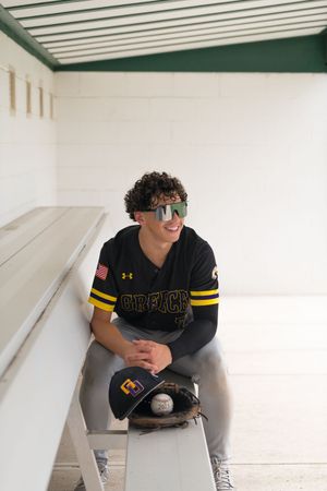 Smiling teen baseball player in Greece uniform sits in dugout wearing sport sunglasses with glove and ball.