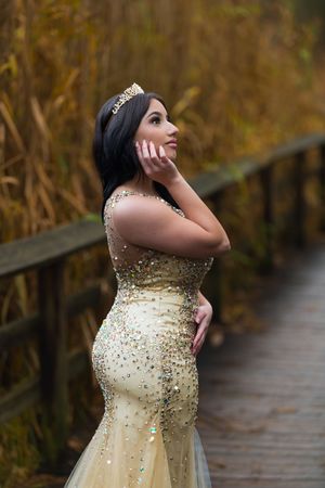 Woman in gold sequin mermaid gown and tiara posing on wooden bridge in autumn setting.