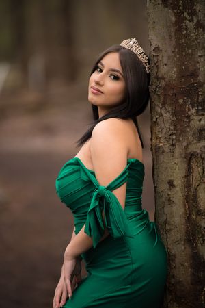 Brunette woman wearing green strapless mermaid gown and gold tiara leaning against tree outdoors.