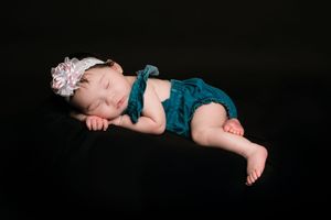 Sleeping newborn baby in teal romper with floral headband on dark background.