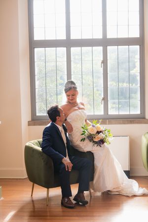 Bride and groom share a romantic moment by a large window, she holds a bouquet while he sits in a green chair.