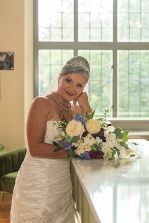 Elegant bride in a strapless white gown and tiara leans on a counter holding a beautiful floral bouquet.