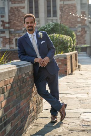 Dapper groom in a navy blue suit with boutonniere leans against a brick wall at an outdoor wedding venue.