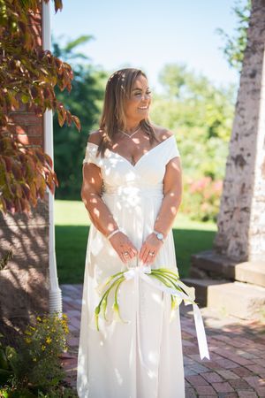 Smiling bride in white off-shoulder gown holding calla lily bouquet outdoors in sunny garden setting.