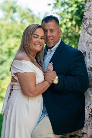 Romantic couple embracing by a tree in lush green garden, woman in white dress, man in navy suit.