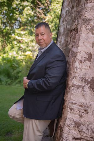 Professional man in navy blazer leaning against tree trunk in lush green garden setting.