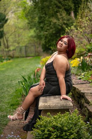 Young woman with red hair in black dress poses on garden bench among spring flowers.