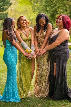 Four young women in colorful prom gowns stack hands together laughing in green park.