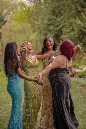 Four laughing young women in elegant prom dresses celebrate together outdoors in lush garden.