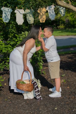 A pregnant woman in white bends down affectionately toward a young boy in a sunny garden setting.